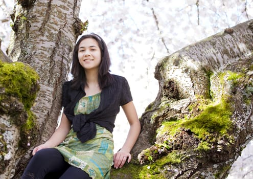 Young biracial teen girl sitting on moss covered branches of large flowering cherry tree