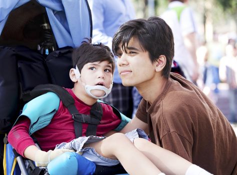 Big brother taking care of disabled little boy in wheelchair outdoors. Child has cerebral palsy.