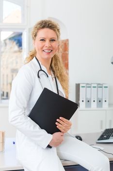 Young female doctor sitting on desk in front of clinic window with test result file or dossier