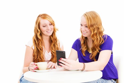 two smiling redhead women sitting at a coffee table looking at a smartphone on white background