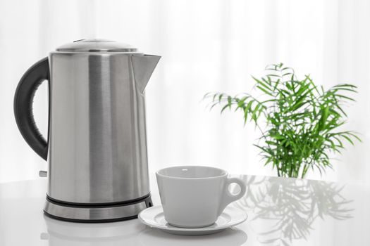 White cup and electric kettle on the table, with green plant in the background.