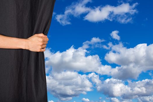 Hand opening black curtain with blue sky and clouds behind it.