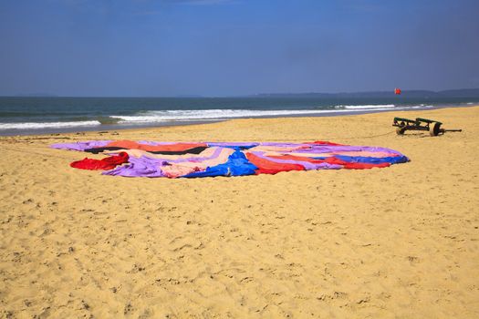Generic horizontal beach and ocean landscape of a parachute laid flat on the sandy Uttorda Beach at Goa, drying in the hot Indian sun. Red tide flag and trolley with waves splashing on the shoreline