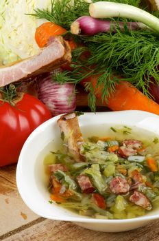 Gourmet Vegetable Soup with Smoked Pork Ribs in White Bowl and Ingredients with Cabbage, Tomatoes, Spring Onion, Leek, Carrot, Garlic and Greens on Wooden background