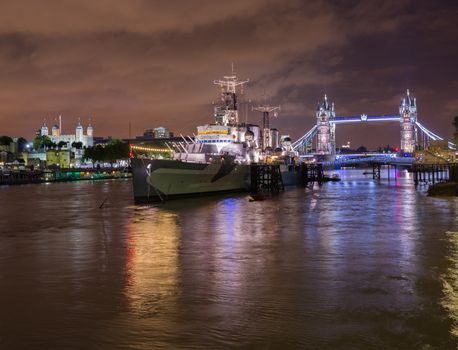 Floodlit HMS Belfast, Tower Bridge and Tower of London on River Thames with reflections in the water