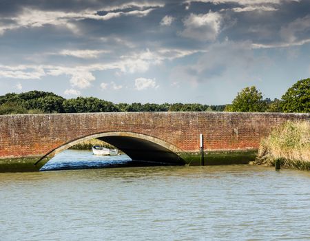 Road arch over Norfolk Broads river with small white rowing boat moored by the bank