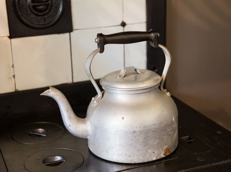 Old fashioned metal kettle sitting on cast iron black cooker hob with tiled surround