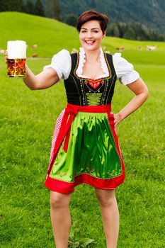 Happy young woman in a dirndl with a large glass tankard of frothy beer raised in her hand in a toast as she stands in a lush green mountain pasture
