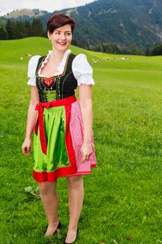 Beautiful stylish young Bavarian woman in a colourful embroidered red and green dirndl standing smiling happily in a lush green mountain meadow