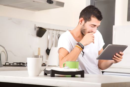 A young male sitting in the kitchen with a Tablet PC.