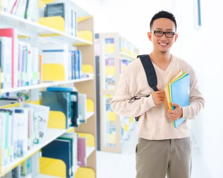 Good looking Southeast Asian adult student in casual wear with school bag carrying text books standing inside school library building.