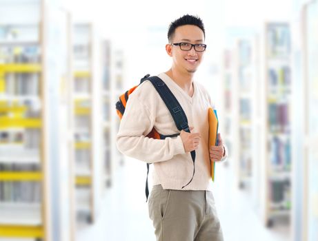 Southeast Asian adult student in casual wear with school bag carrying text books standing inside school library building.