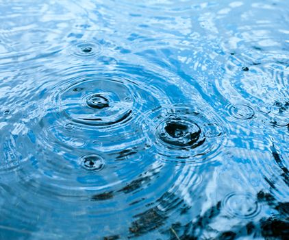 Rain drops rippling in a puddle with blue sky reflection