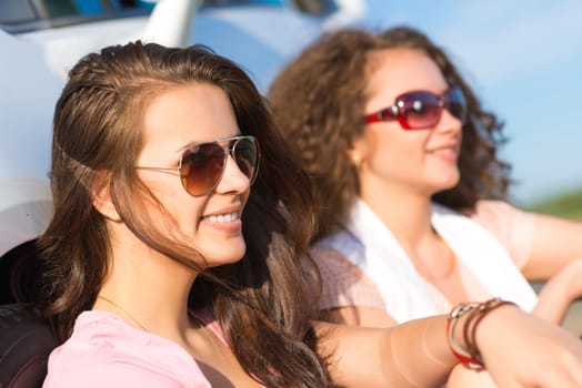 Two attractive young women wearing sunglasses, sitting next to the car