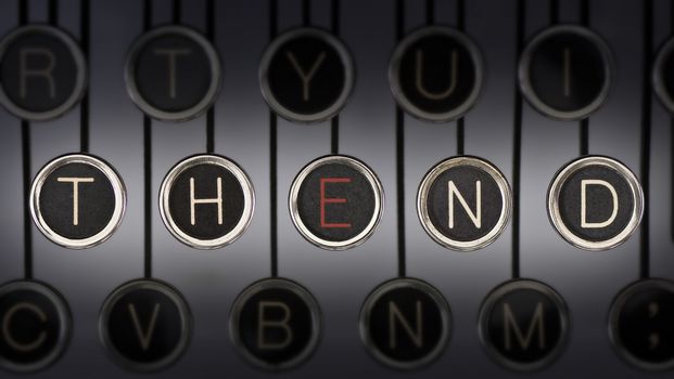 Close up of old manual typewriter keyboard with scratched chrome keys that spell out "THE END". Both words share the letter, 'E', highlighted in red.  Lighting and focus are centered on "THE END" 

