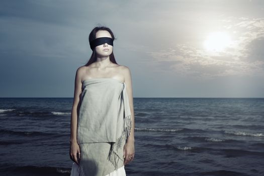 Lonely woman with blindfold listening waves of the sea during sunset. Special colors and processing in artistic manner