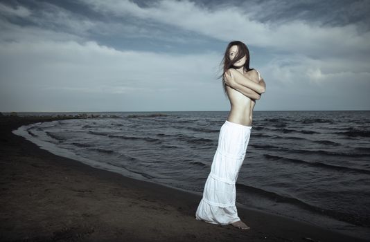 Single topless lady in the white shirt standing near the sea. Hairs are blown by the wind. Artistic colors and darkness added