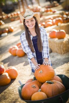 Preteen Girl Wearing Cowboy Hat Playing with a Wheelbarrow at the Pumpkin Patch in a Rustic Country Setting.
