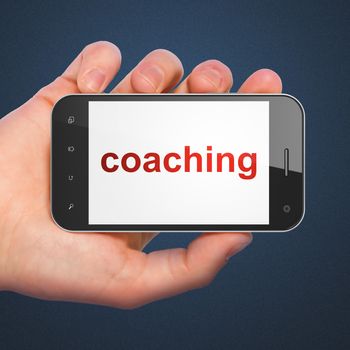 Education concept: hand holding smartphone with word Coaching on display. Generic mobile smart phone in hand on Dark Blue background.