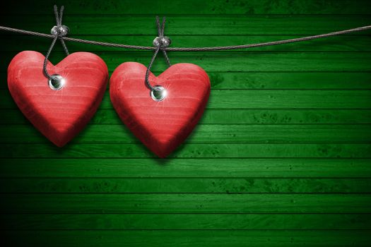 Two handmade red wooden hearts hanging on a steel cable on green wooden background