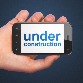 SEO web design concept: hand holding smartphone with word Under Construction on display. Mobile smart phone in hand on Blue background.