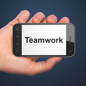 Business concept: hand holding smartphone with word Teamwork on display. Generic mobile smart phone in hand on Dark Blue background.