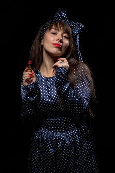 Elegant sensual woman with long brunette hair wearing stylish polka dot evening dress applying red lipstick to her lips against a dark background