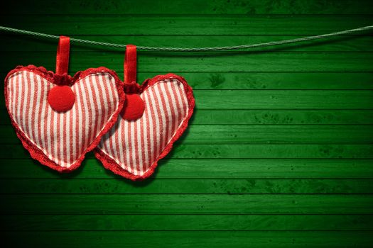 Two handmade red and white cloth hearts hanging on a steel cable on green wooden background