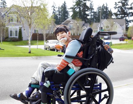 Happy disabled six year old boy waiting on sidewalk in wheelchair. Child has cerebral palsy