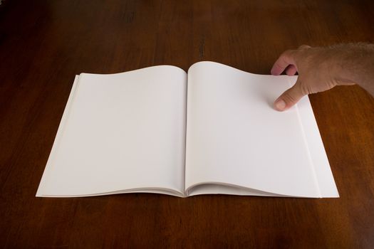 Open blank book or magazine with white paper. Insert your own design. There is a hand located at the top left of the open book / magazine, ready to turn the page. The open book / magazine is sitting on a wooden tabletop.