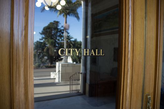 A door leading inside of a southern California City Hall building. Palm trees and the street are visible in the reflection of the glass door.