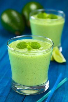 Healthy refreshing green smoothie made of fresh avocado, lime and yoghurt garnished with mint and served in glasses on blue wood (Selective Focus, Focus on the big mint leaves on the drink)