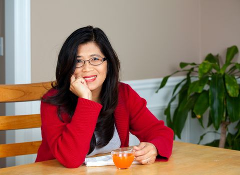 Asian woman in red sweater relaxing at dining table with glass of juice, smiling