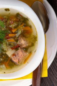 Tasty Homemade Vegetable Soup with Boiled Beef and Greens in White Bowl with Spoon on Yellow Napkin. Top View