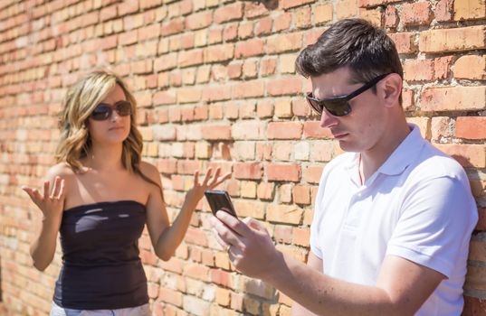 Angry man focused looking phone and displeased girl up her hands in front of wall background