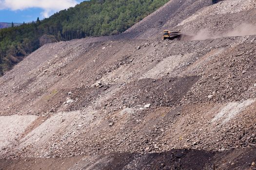 Big mining truck on haul road throwing up dust off its rough surface at steep mountain side with tailings