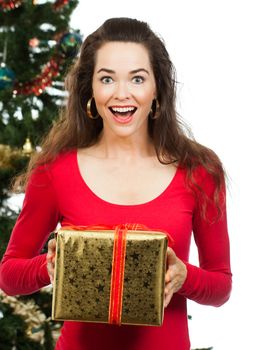 Surprised beautiful woman holding a Christmas gift in front of a Christmas tree and looking at camera. Isolated on white.