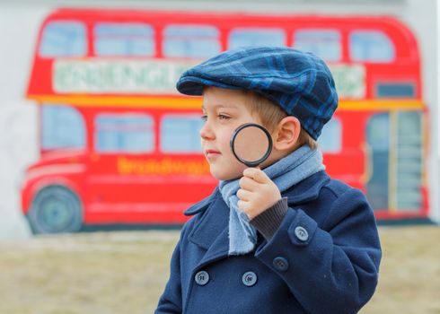 Little detective. Funny boy looking through magnifying glass near english schoolbus