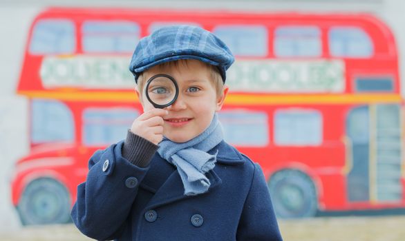 Little detective. Funny boy looking through magnifying glass near english schoolbus