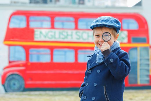 Little detective. Funny boy looking through magnifying glass near english schoolbus