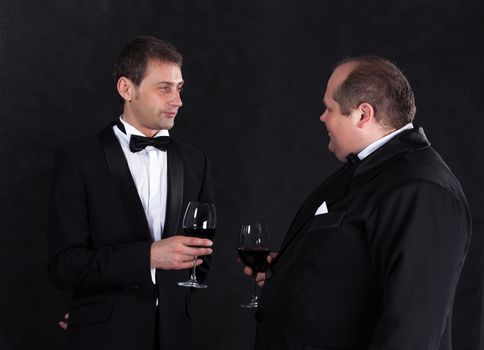 Two stylish businessman in tuxedos with glasses of red wine, on black background