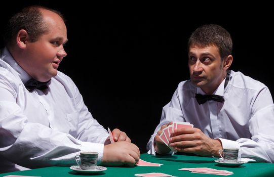 Two gentlemen in white shirts, playing cards, on black background