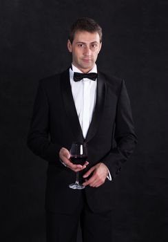 Stylish man in elegant black tuxedo with glass red wine, on black background