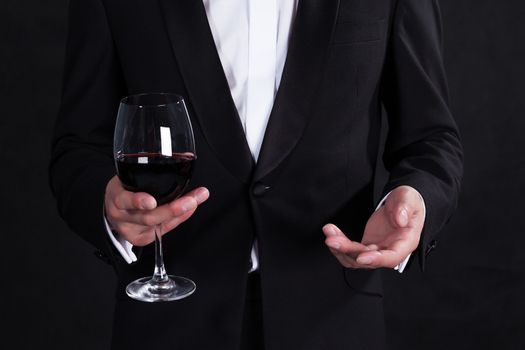 Fragment of stylish man in elegant black tuxedo with glass red wine, on black background