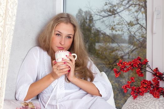 Portrait of charming girl in sweater holding cup and looking through window