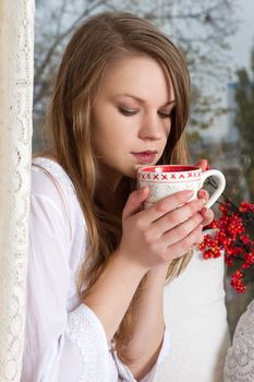 Portrait of charming girl in sweater holding cup and looking through window