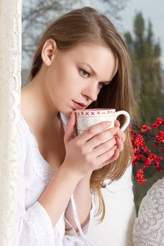 Portrait of charming girl in sweater holding cup and looking through window