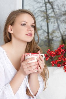 Portrait of charming girl in sweater holding cup and looking through window