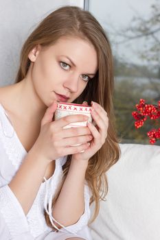 Portrait of charming girl in sweater holding cup and looking through window