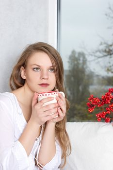 Portrait of charming girl in sweater holding cup and looking through window